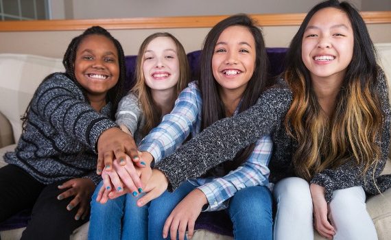 Diverse group of teen girls sitting on a couch touching hands.