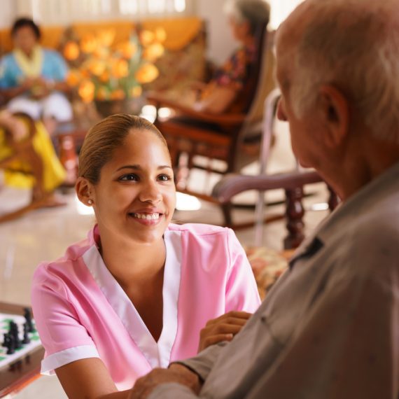 young attractive hispanic woman working as nurse takes care of a senior man on wheelchair. She talks with him then goes away to help other patients