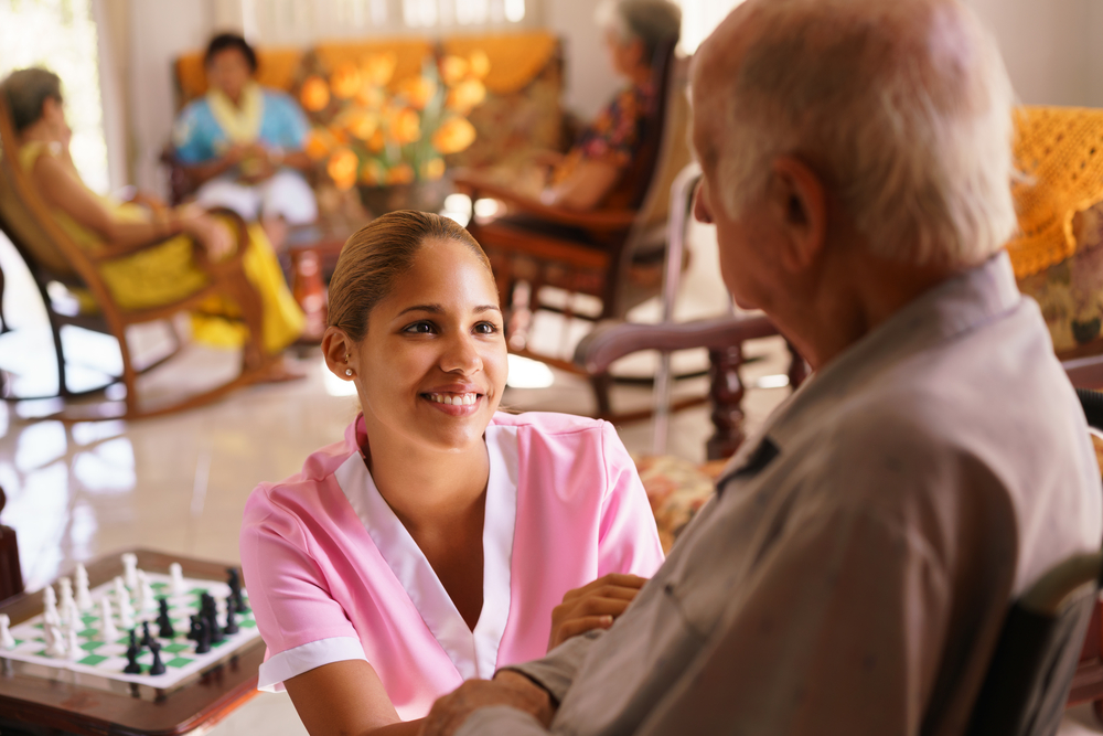 young attractive hispanic woman working as nurse takes care of a senior man on wheelchair. She talks with him then goes away to help other patients