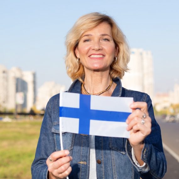 Happy mature woman with flag of Finland standing against the background of a city street and blue sky.