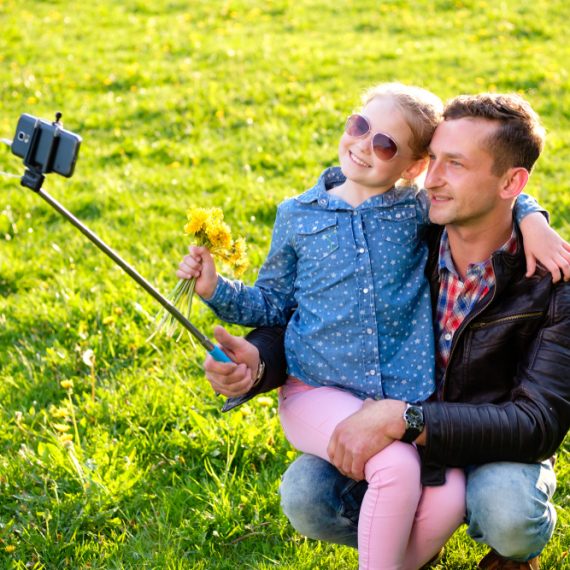 Happy Dad and little daughter doing picture by smartphone use selfie stick in summer park.