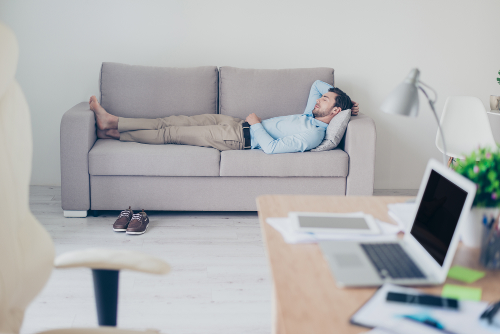 Concept of necessity of having a rest while working. Tired exhausted manager clothed in formal-wear is sleeping on a sofa in his modern office