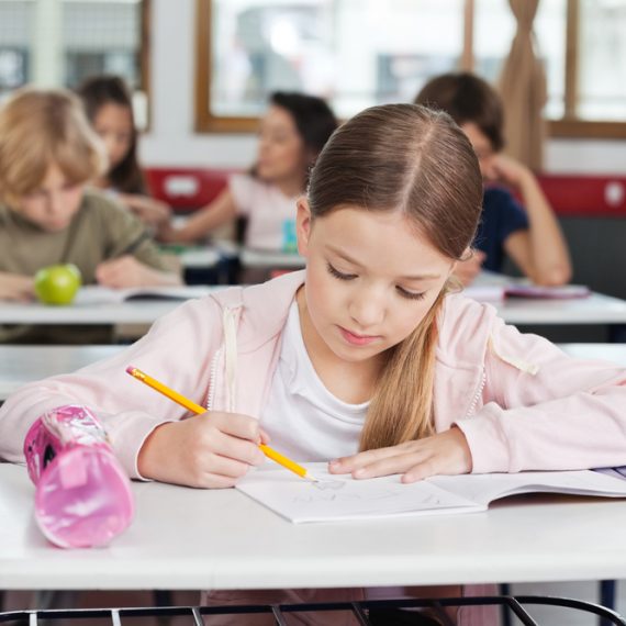 Cute little schoolgirl drawing in book with classmates in background at classroom