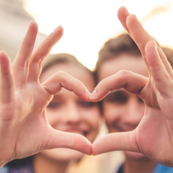 Beautiful young couple is making heart of fingers, looking through it and smiling, hands in focus, close-up