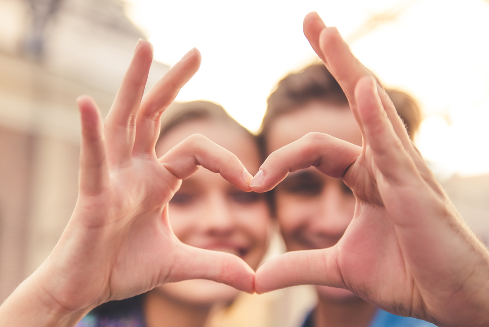 Beautiful young couple is making heart of fingers, looking through it and smiling, hands in focus, close-up