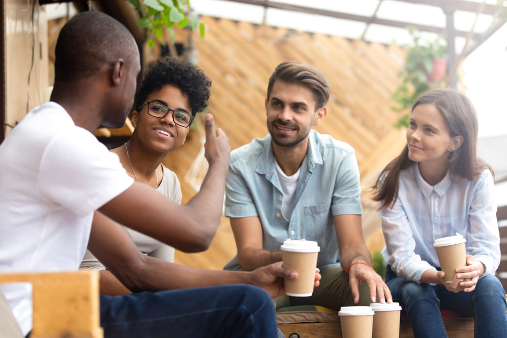 African American man showing thumb up, telling story to interested attentive friends, sitting, drinking coffee in cafe together, smiling multiethnic friends discussing, talking, chatting