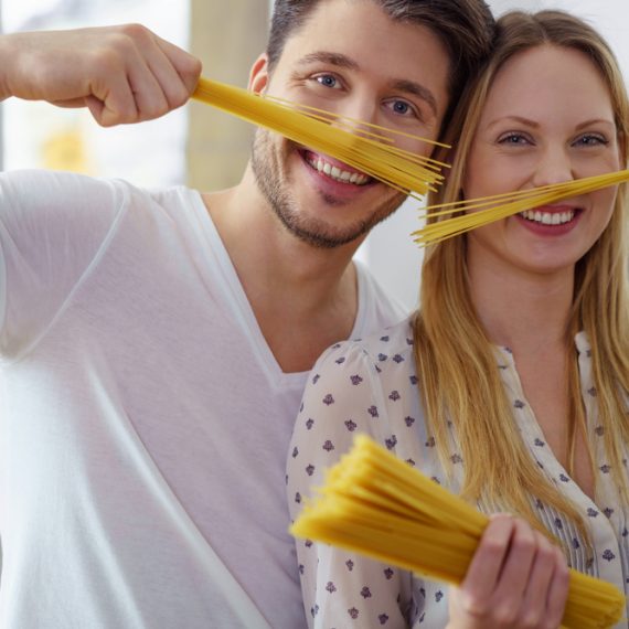 Silly young husband and wife in their kitchen playing with straight uncooked pasta noodles near their puckered faces