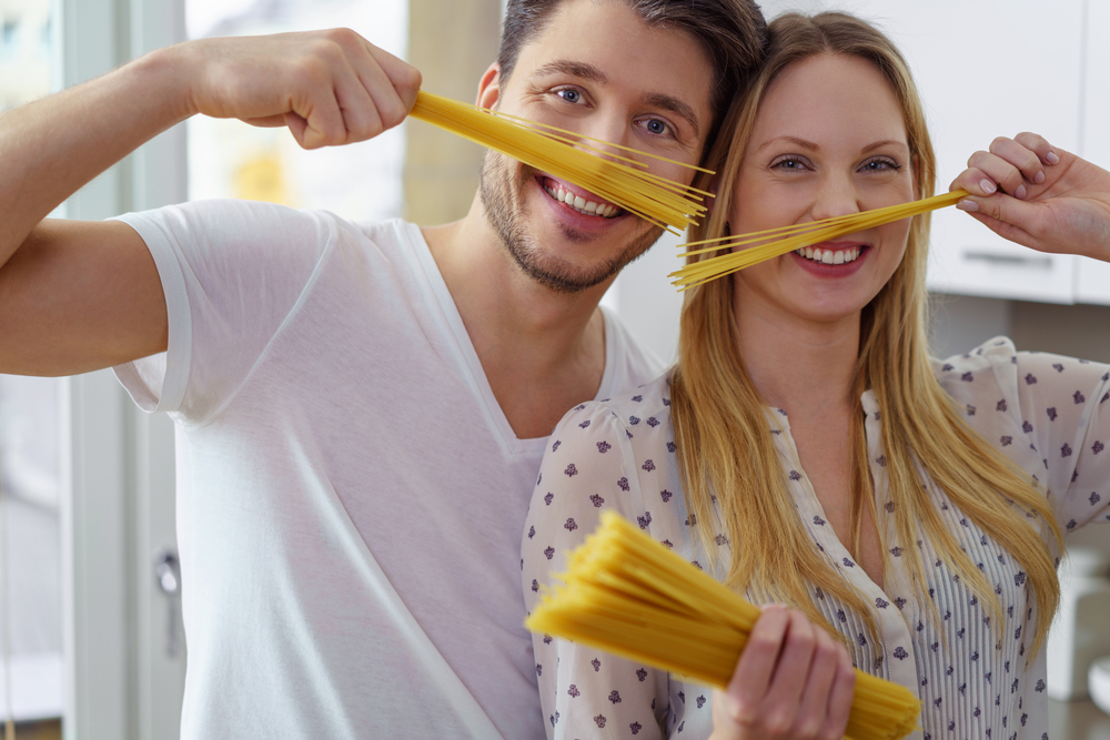 Silly young husband and wife in their kitchen playing with straight uncooked pasta noodles near their puckered faces