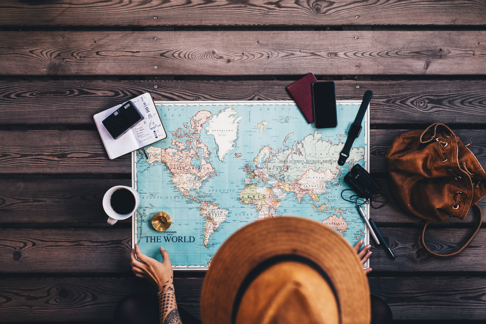 Young woman planning vacation using world map and compass along with other travel accessories. Tourist wearing brown hat looking at the world map.