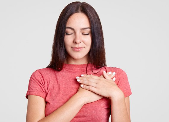 Calm brunette female with closed eyes, keeps both palms on heart, feels gratitude, being touched by something, dressed in casual pink t shirt, isolated over white background.