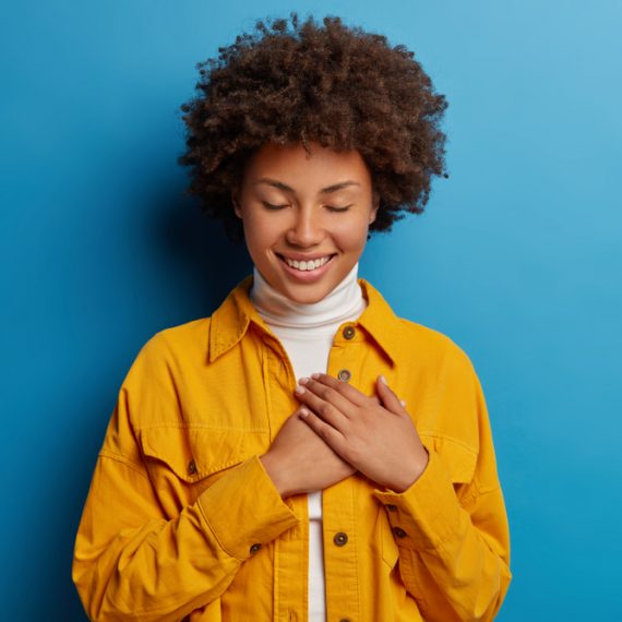 Tender feminine woman keeps hands on heart, has grateful look, appreciates effort, has eyes closed, wears yellow shirt, isolated over blue background, receives touching compliment, smiles pleasantly