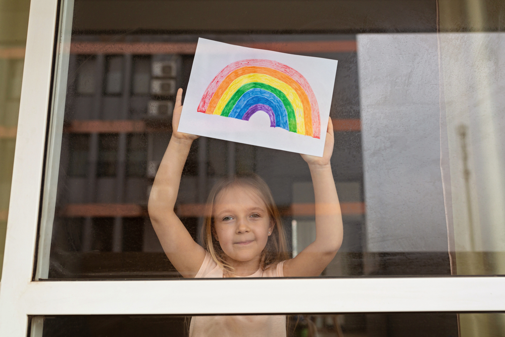 Kid painting rainbow during Covid-19 quarantine at home. Girl near window. Stay at home Social media campaign for coronavirus prevention, let's all be well, hope during coronavirus pandemic concept