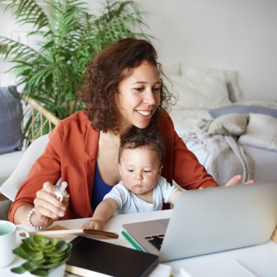 Attractive young dark skinned woman working at desk at home using laptop, holding baby on her lap. Portrait of smiling mother writing post on moms blog while her infant son playing with toy s