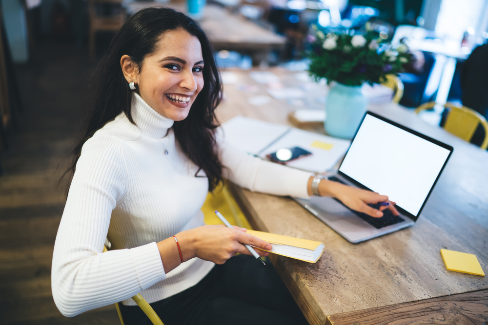 Happy smiling lovely female in casual wear with notepad and pen typing using laptop working on project sitting at table in cafeteria