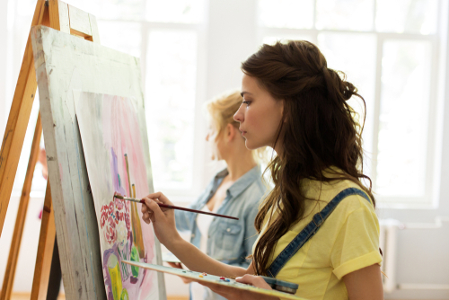 art school, creativity and people concept - student girl or young woman artist with easel, palette and paint brush painting still life picture at studio