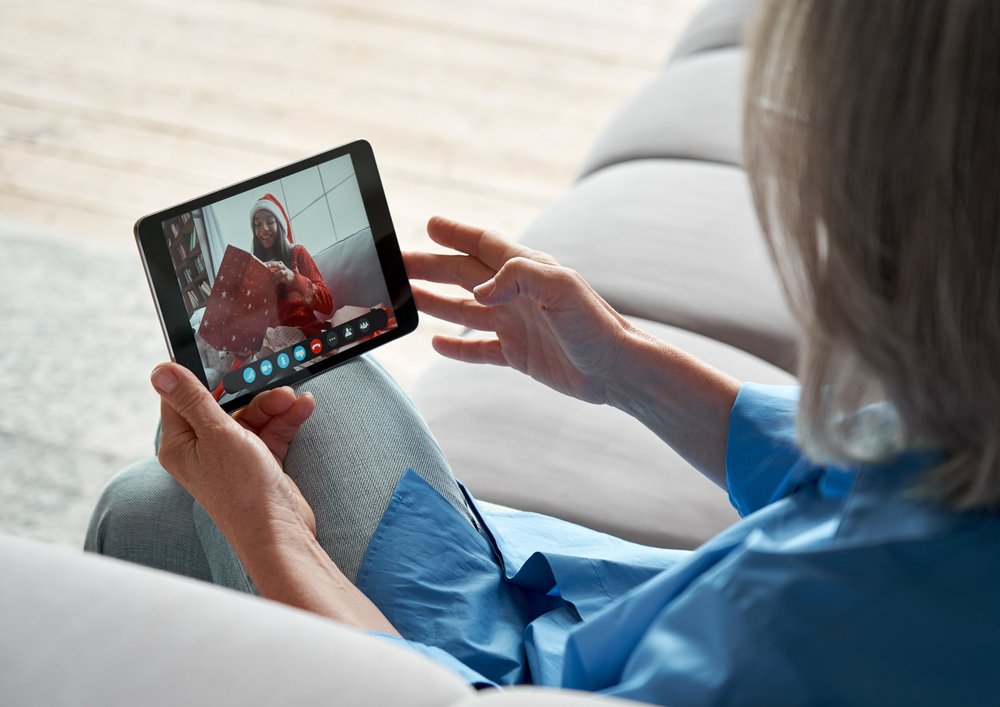 Over shoulder view of old senior grandmother holding digital tablet computer in hands video conference calling granddaughter opening Christmas gift during distance virtual family online chat meeting.