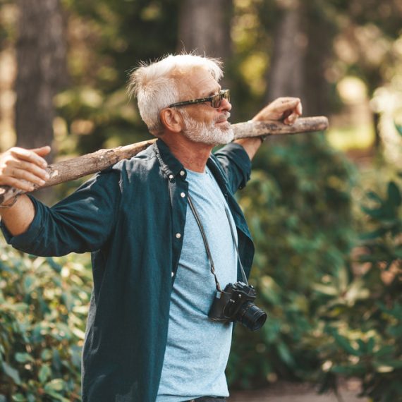 A mature bearded grandfather holds a stick on his shoulders. A pensioner man enjoys life, walking in the woods. Outdoor activities in old age.