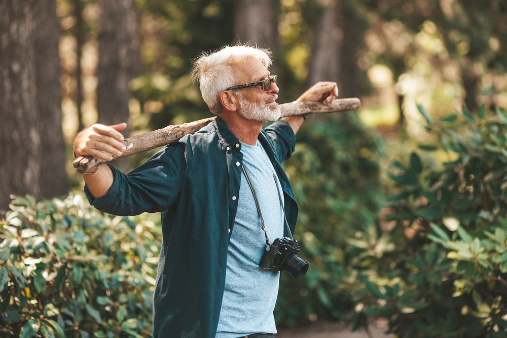 A mature bearded grandfather holds a stick on his shoulders. A pensioner man enjoys life, walking in the woods. Outdoor activities in old age.