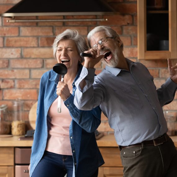 Happy senior husband and wife have fun sing in kitchen appliances cooking together at home.