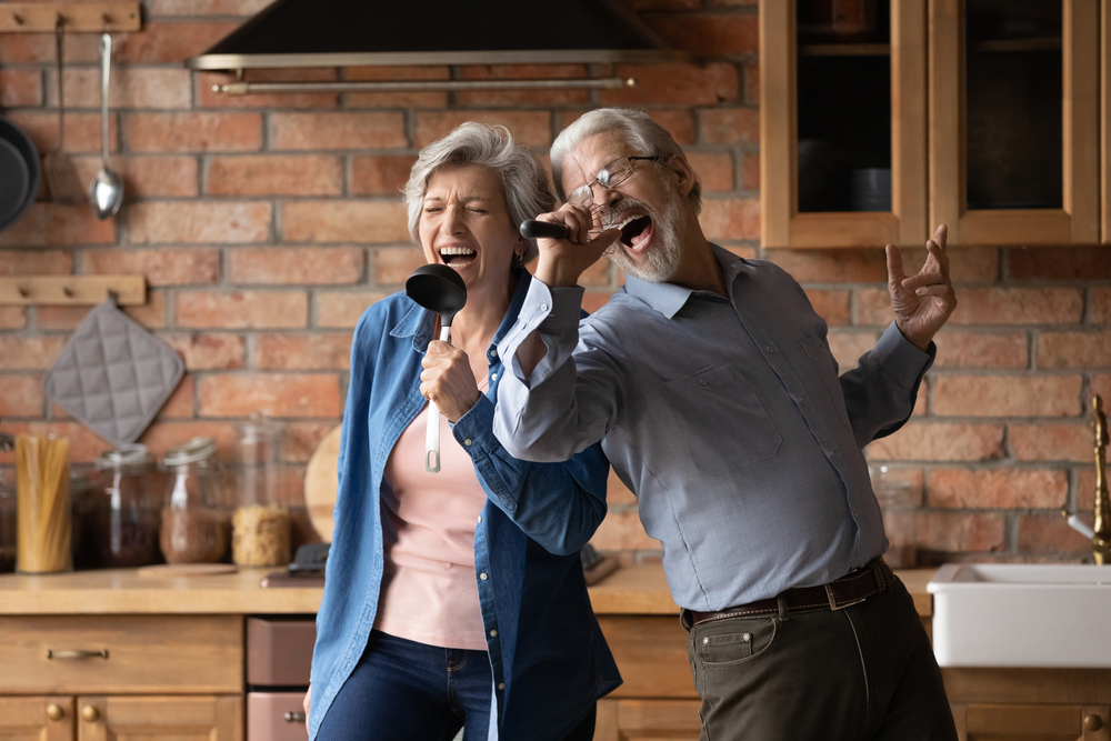 Happy senior husband and wife have fun sing in kitchen appliances cooking together at home.