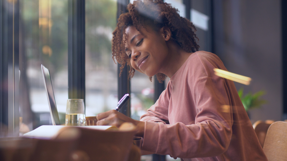 Side view of one happy young African woman writing indoor, Pretty Black college student study with laptop by the window doing homework writing letter