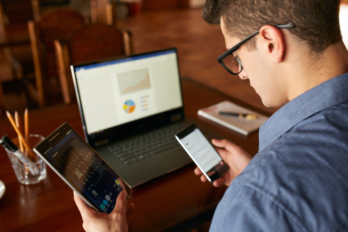 Attractive man in glasses working with multiple electronic internet devices. Freelancer businessman has laptop and smartphone in hands and laptop on table with charts on screen.